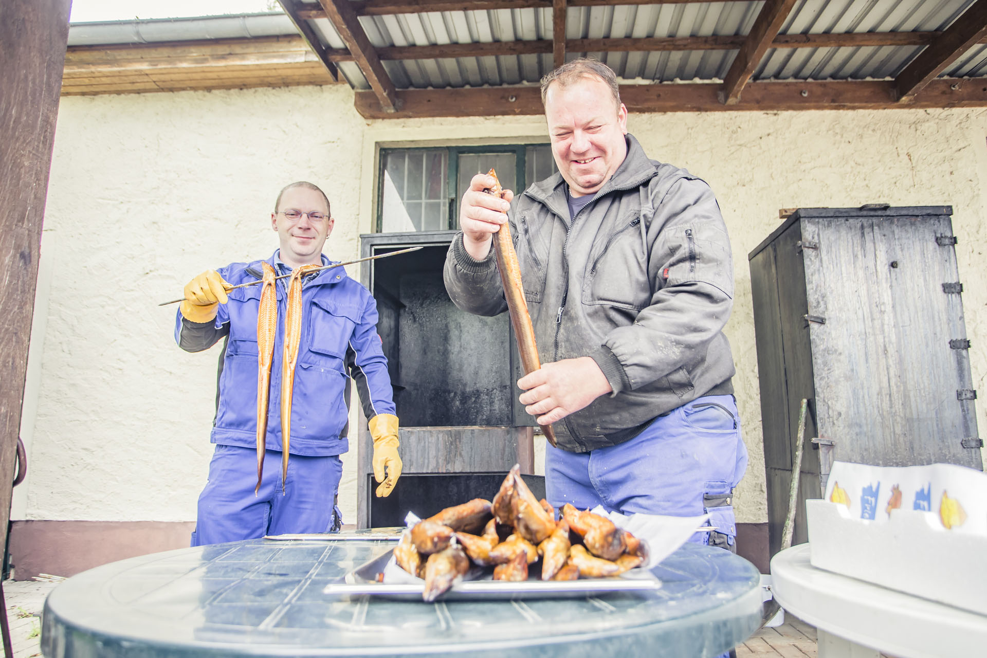Ein Mann legt die geräucherten Aale auf anderen ab, während der andere Mann den Spieß mit den restlichen geräucherten Aalen hält